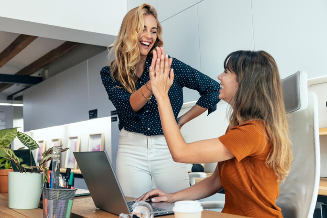 Two smiling coworkers giving a high five at a desk in a modern office, celebrating a successful moment.